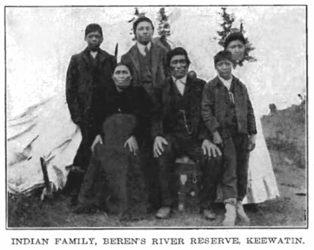 Black and white photo of an Indigenous family. Three boys in suits and a girl i na white dress are standing. Sitting in front of them are two parents, a mother and a father. She is wearing a dark shirt and skirt. He is wearing a suit. 